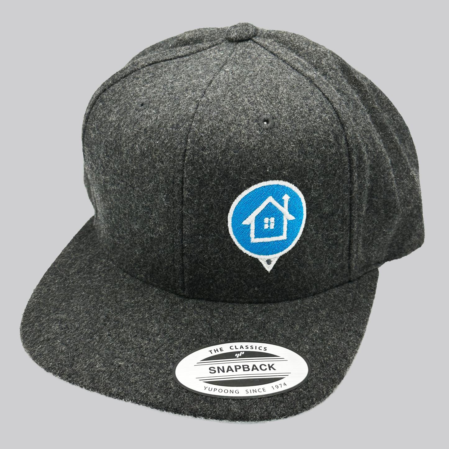 A dark heather gray wool baseball cap with a high profile and flat bill. The front left of the cap features an embroidered HomeValue.com logo – a light blue map marker circle with white edging and a white embroidered house inside the circle. The underside of the cap’s visor is green.
