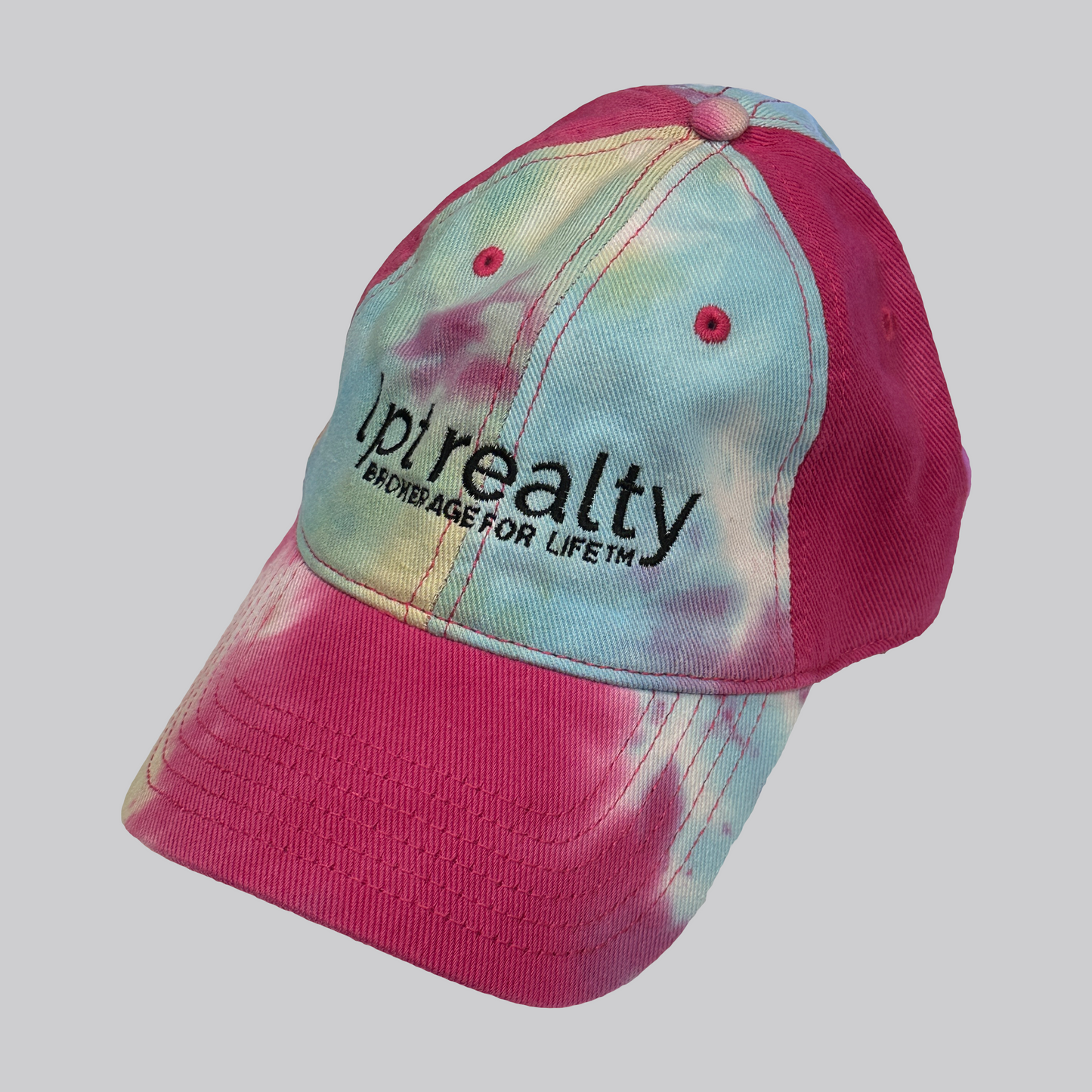 A bright, colorful tie-dye baseball cap featuring hot pink stitching and eyelet embroidery. The cap’s text is embroidered in black, with “Lpt Realty” in large, lowercase letters and “Brokerage For Life™” underneath in smaller, all-caps text.