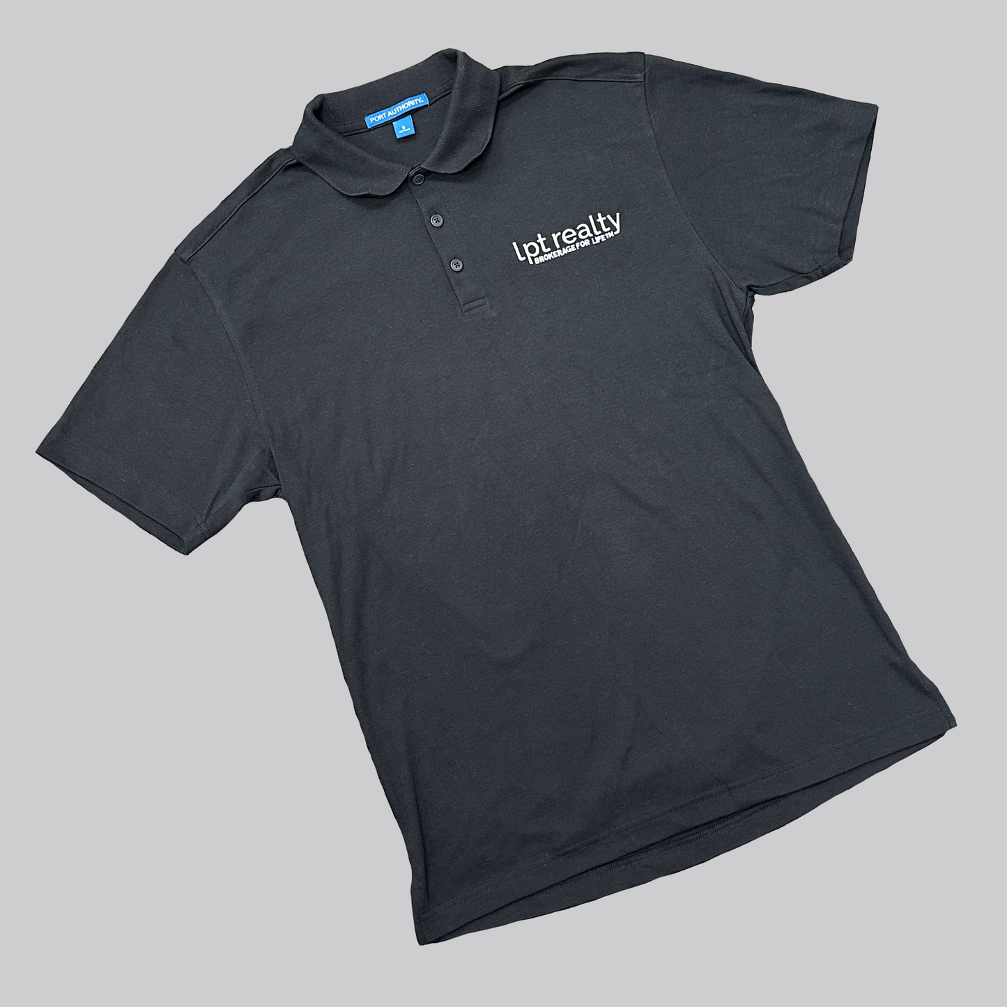 A black polo with black buttons and “lpt realty” embroidered in white in small, lowercase letters on the upper left chest. Below this, in smaller, all-caps text, is the trademarked phrase “Brokerage For Life™“.