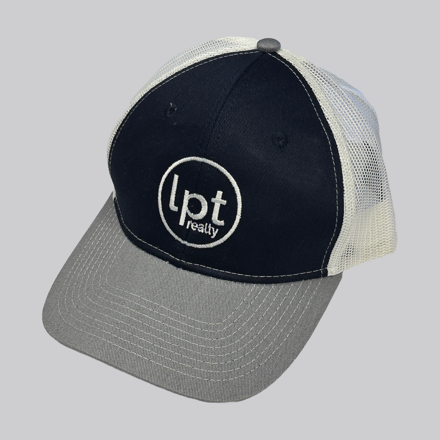 Classic trucker hat design with solid front panels and white mesh back. The front panels are a rich navy blue with matching embroidered eyelets and an embroidered white “Lpt Realty” circle logo at the center. The bill is a medium heather gray with white stitching.