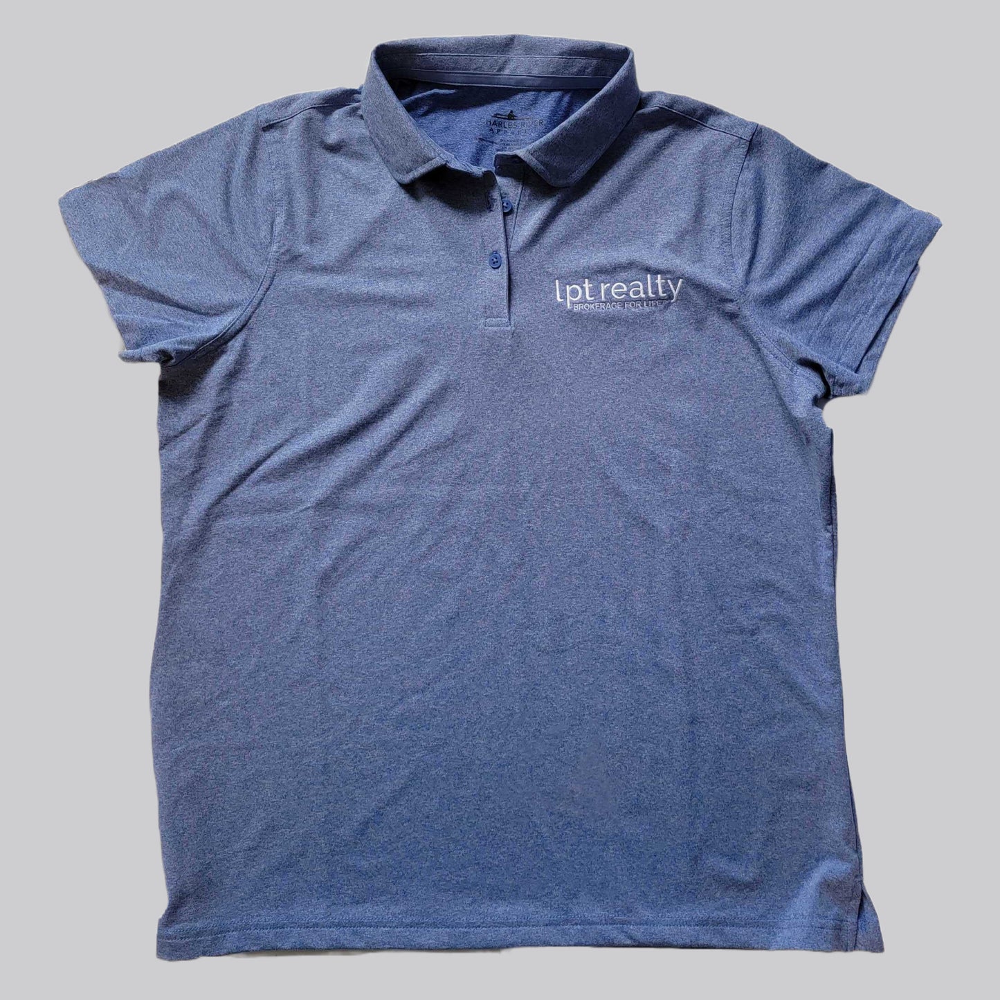 A heathered blue polo shirt with a subtle sheen from the stretch fabric. The upper left chest features “lpt realty” in white, embroidered lowercase letters, with “BROKERAGE FOR LIFE” in smaller all caps beneath it. The shirt’s placket has 3 buttons in matching blue, the collar is the same fabric as the shirt, and the sleeves are open with no cuffs.