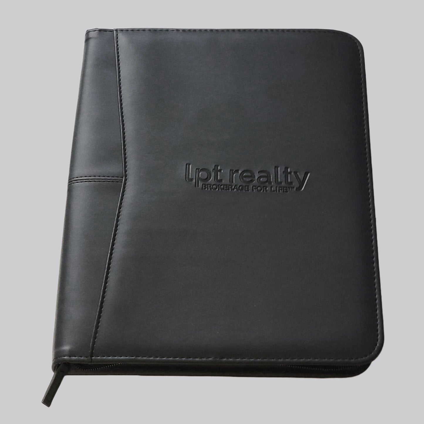 A black faux leather zippered portfolio with “lpt realty” embossed on the front center of the cover, and “BROKERAGE FOR LIFE ™” embossed just below it. Inside, the right side features a white, letter-sized lined notepad. The left side is a panel containing 8 business-card-sized pockets, a larger for a cell phone, and multiple accessory loops. This panel is the outer part of a larger gusseted pocket that can fit a notebook or tablet up to 10 inches.