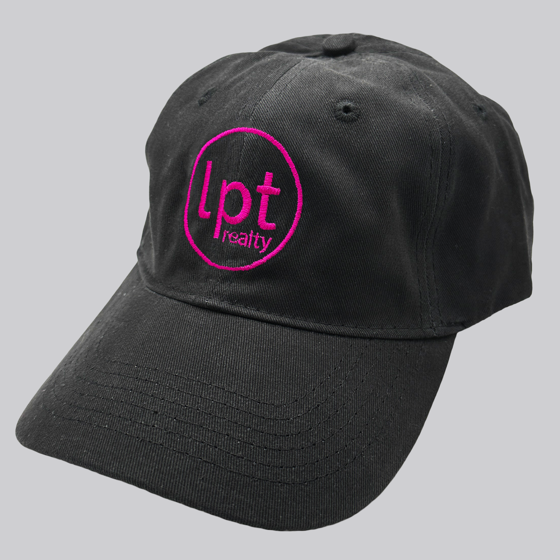 A black twill cap with a relaxed fit, matching stitching, and embroidered eyelets. The front panels feature the “lpt realty” circle logo embroidered in hot pink.  