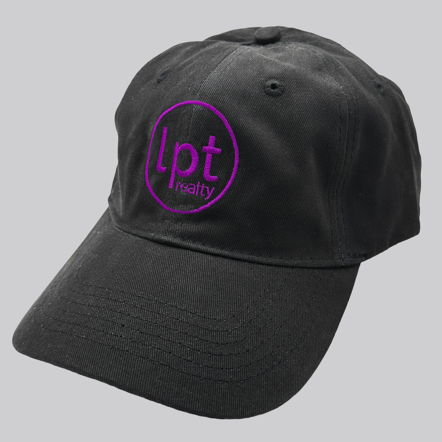 A black twill cap with a relaxed fit, matching stitching, and embroidered eyelets. The front panels feature the “lpt realty” circle logo embroidered in vivid purple. 