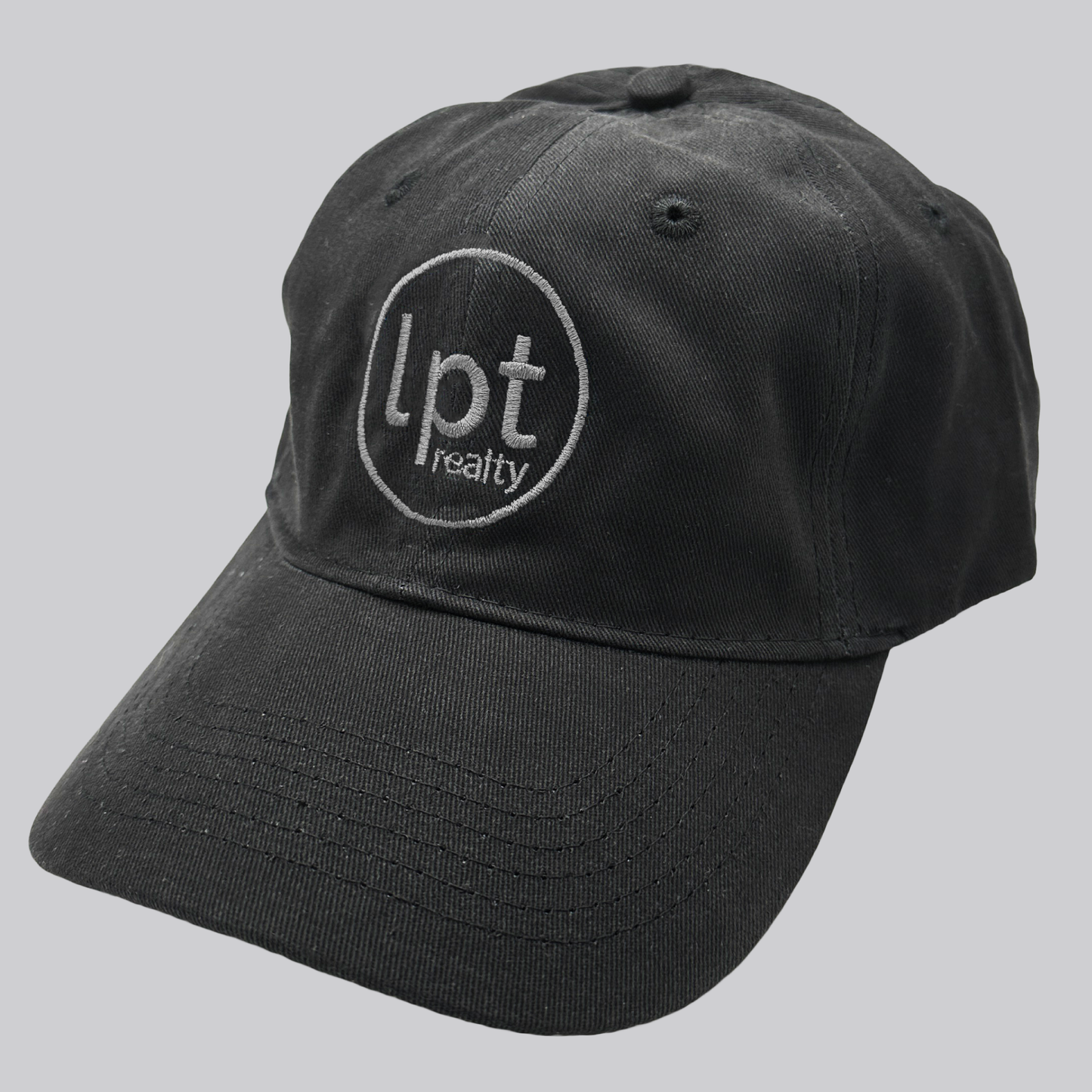 A black twill cap with a relaxed fit, matching stitching, and embroidered eyelets. The front panels feature the “lpt realty” circle logo embroidered in silver. 