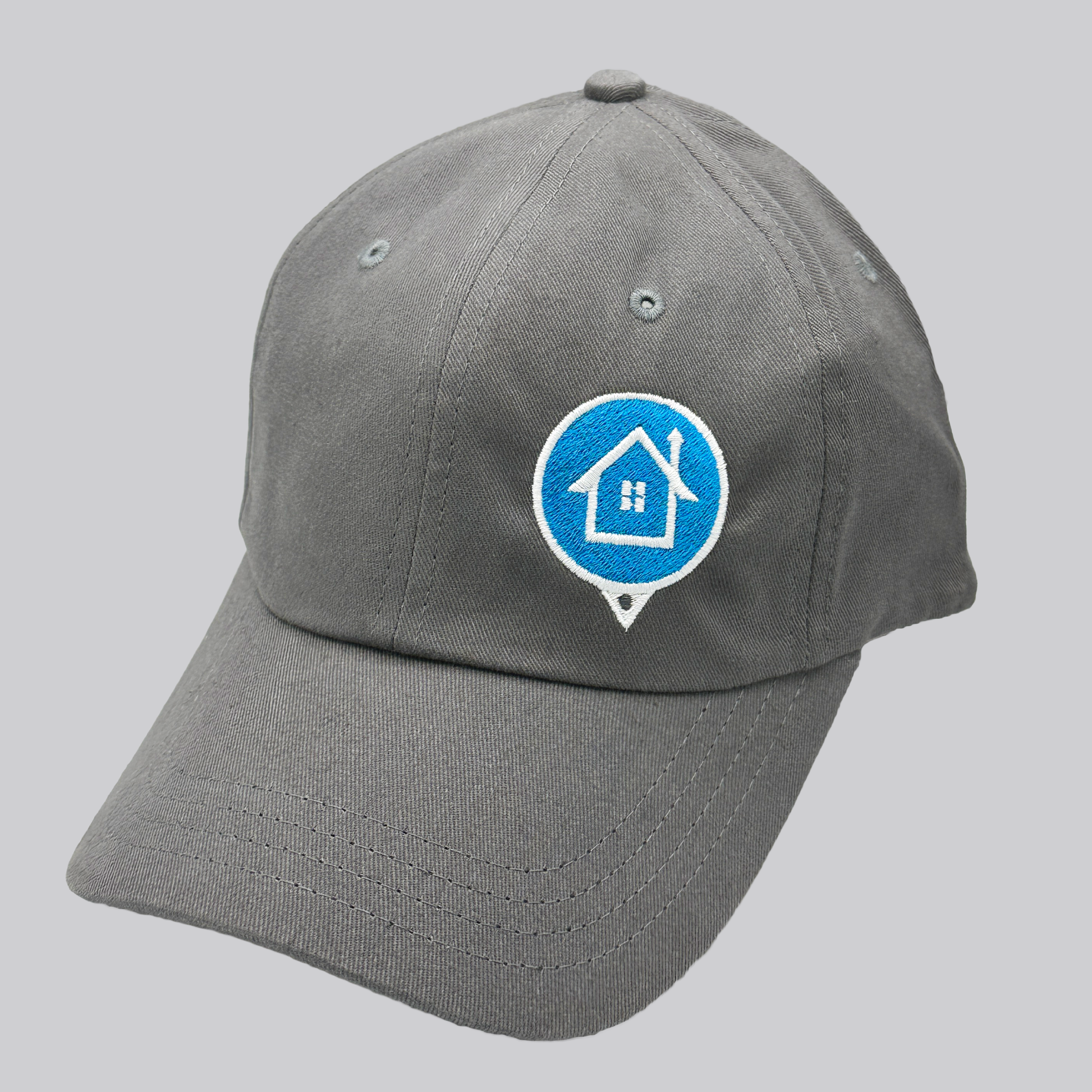 A gray twill cap with a relaxed fit, matching stitching, and embroidered eyelets. The front left panel features an embroidered HomeValue.com logo: a blue circle map marker with a white border and a white house icon in the center. 