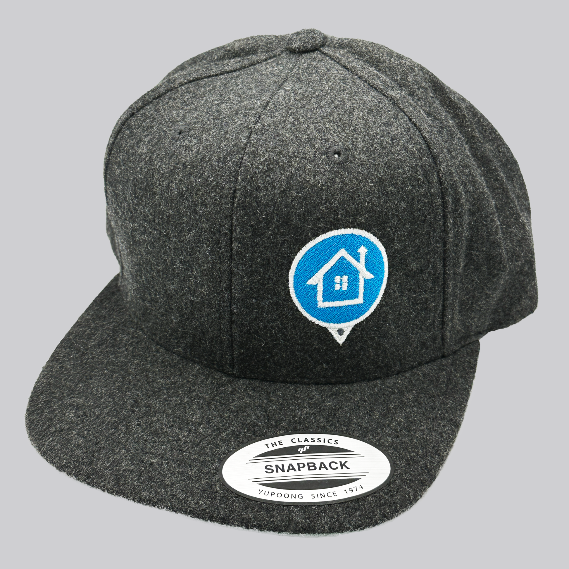 A dark heather gray wool baseball cap with a high profile and flat bill. The front left of the cap features an embroidered HomeValue.com logo – a light blue map marker circle with white edging and a white embroidered house inside the circle. The underside of the cap’s visor is green. 