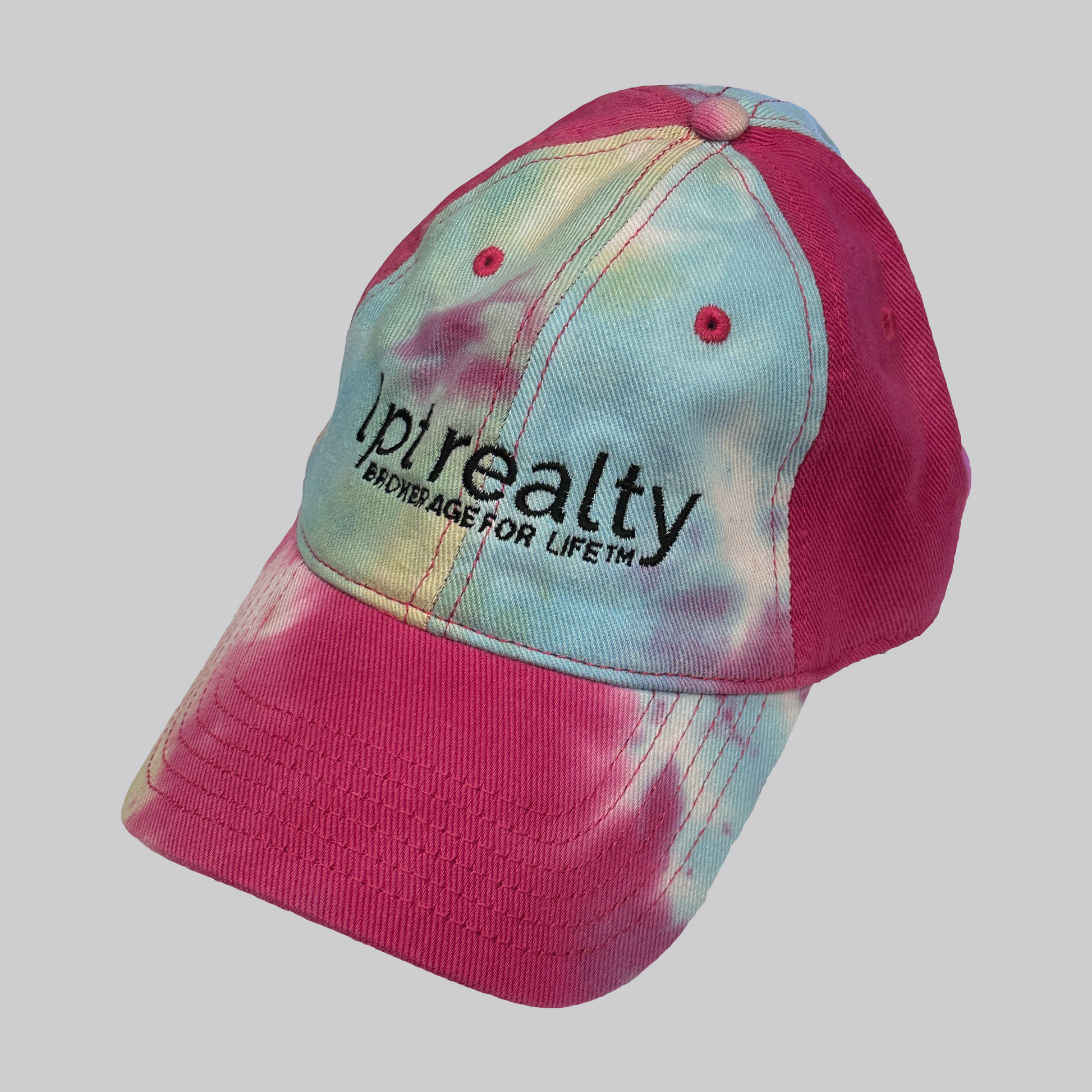 A bright, colorful tie-dye baseball cap featuring hot pink stitching and eyelet embroidery. The cap’s text is embroidered in black, with “Lpt Realty” in large, lowercase letters and “Brokerage For Life™” underneath in smaller, all-caps text. 