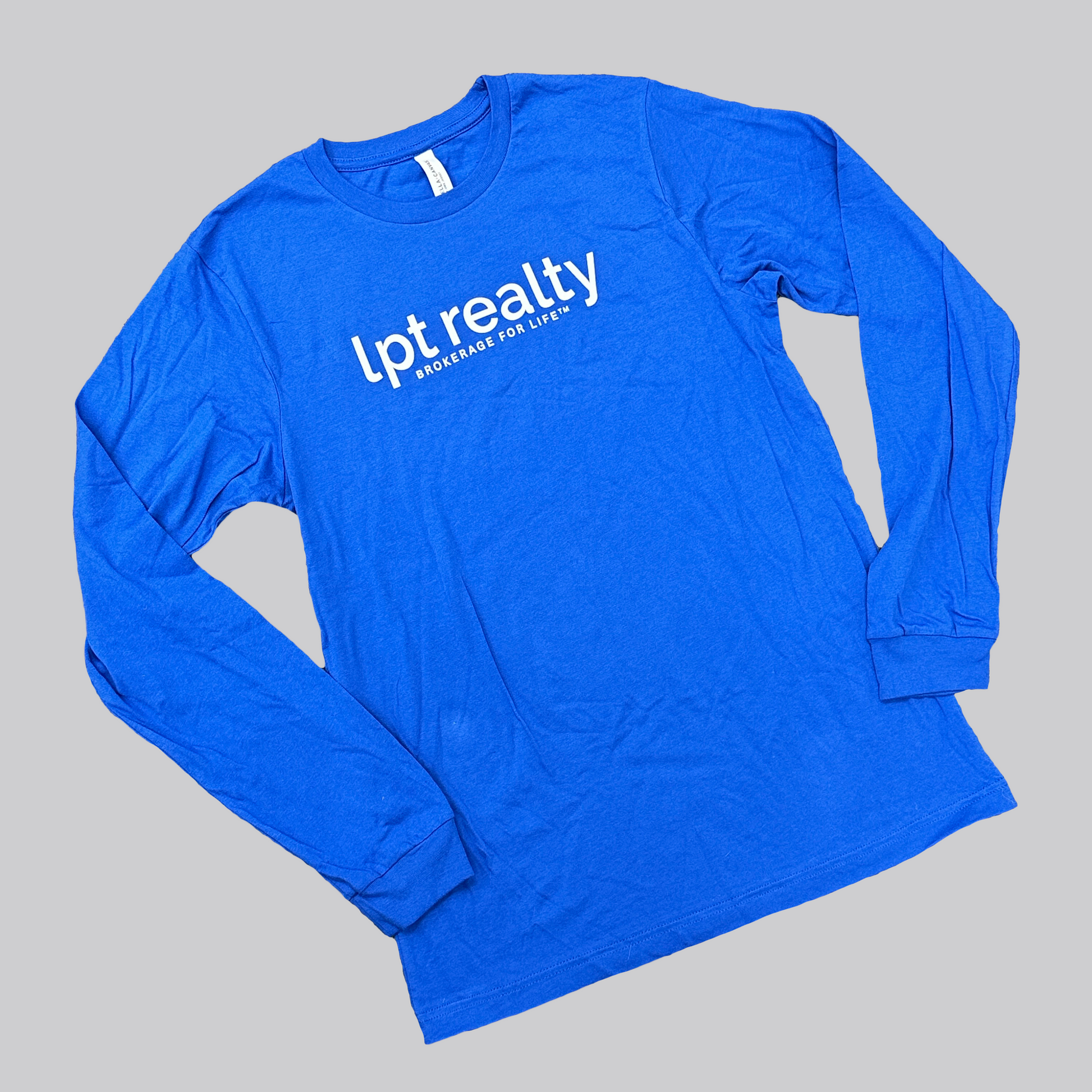 A royal blue long-sleeved t-shirt with ribbed cuffs. The front features white, lowercase text reading 'lpt realty' across the center chest, and beneath it, in smaller all-caps text, "Brokerage For Life™." 