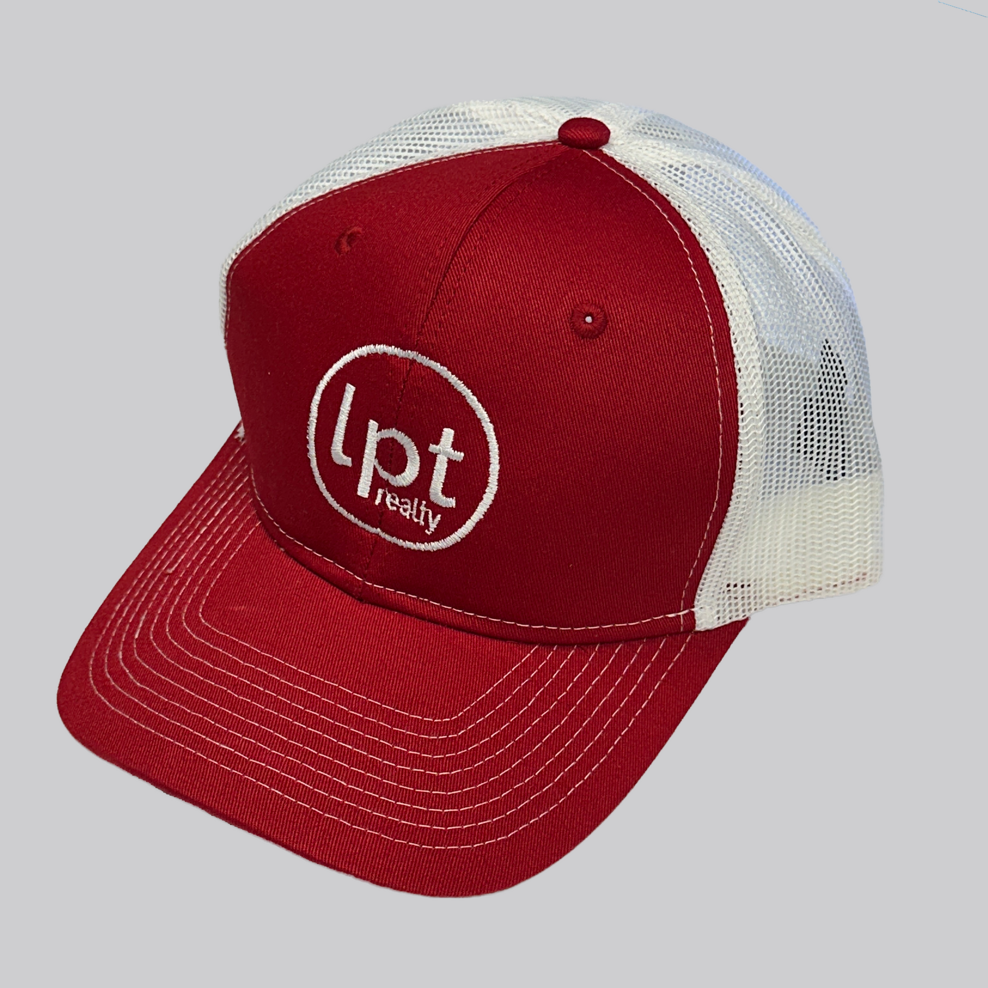 Classic two-toned trucker hat design with solid front panels and white mesh back. The front panels and bill are a solid flame red with matching embroidered eyelets, an embroidered white “Lpt Realty” circle logo at the center, and white stitching on the bill.  