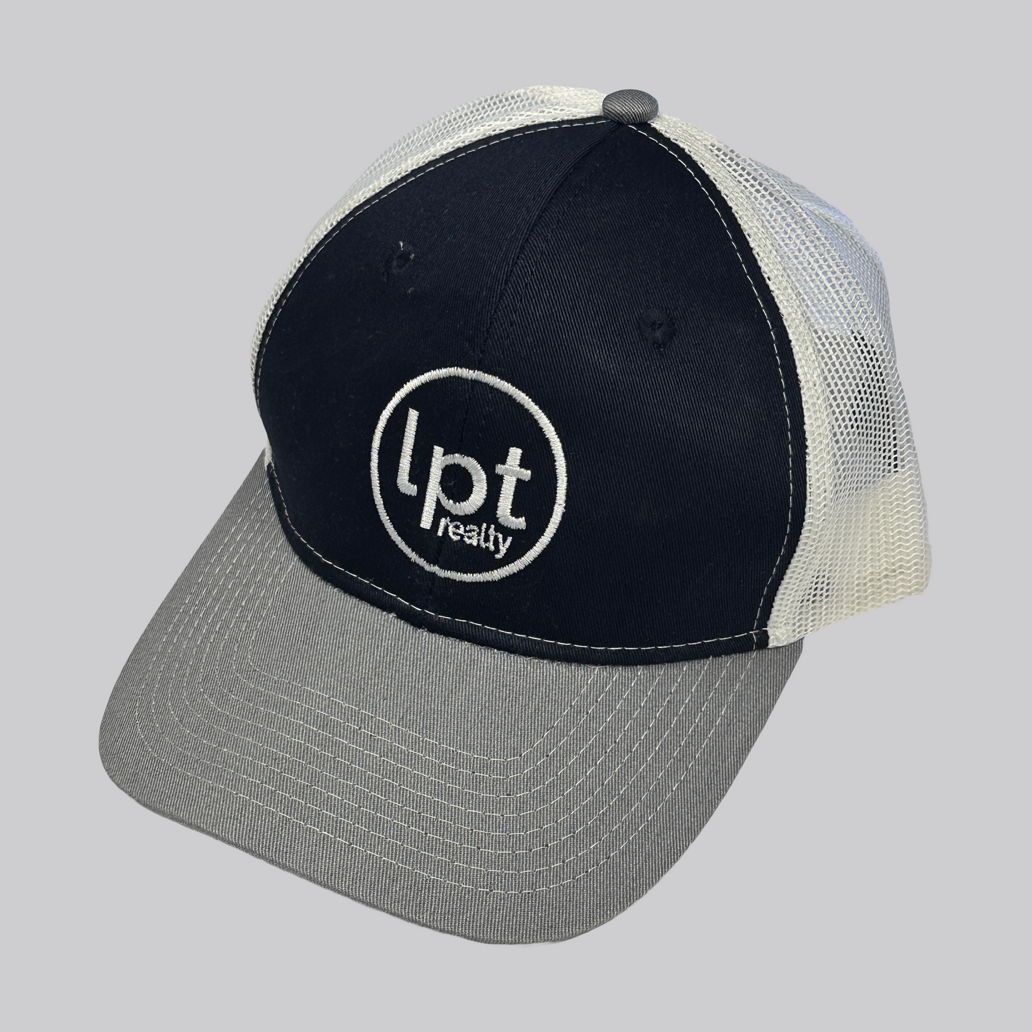 Classic trucker hat design with solid front panels and white mesh back. The front panels are a rich navy blue with matching embroidered eyelets and an embroidered white “Lpt Realty” circle logo at the center. The bill is a medium heather gray with white stitching. 