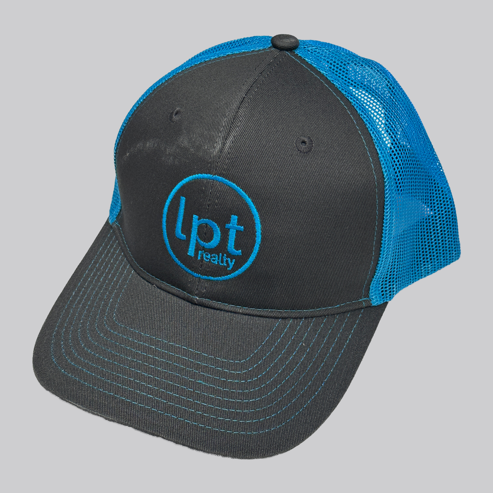 Classic two-toned trucker hat design with solid front panels and neon blue mesh back. The front panels and bill are a solid graphite gray with matching embroidered eyelets, an embroidered neon blue “Lpt Realty” circle logo at the center, and neon blue stitching on the bill. 