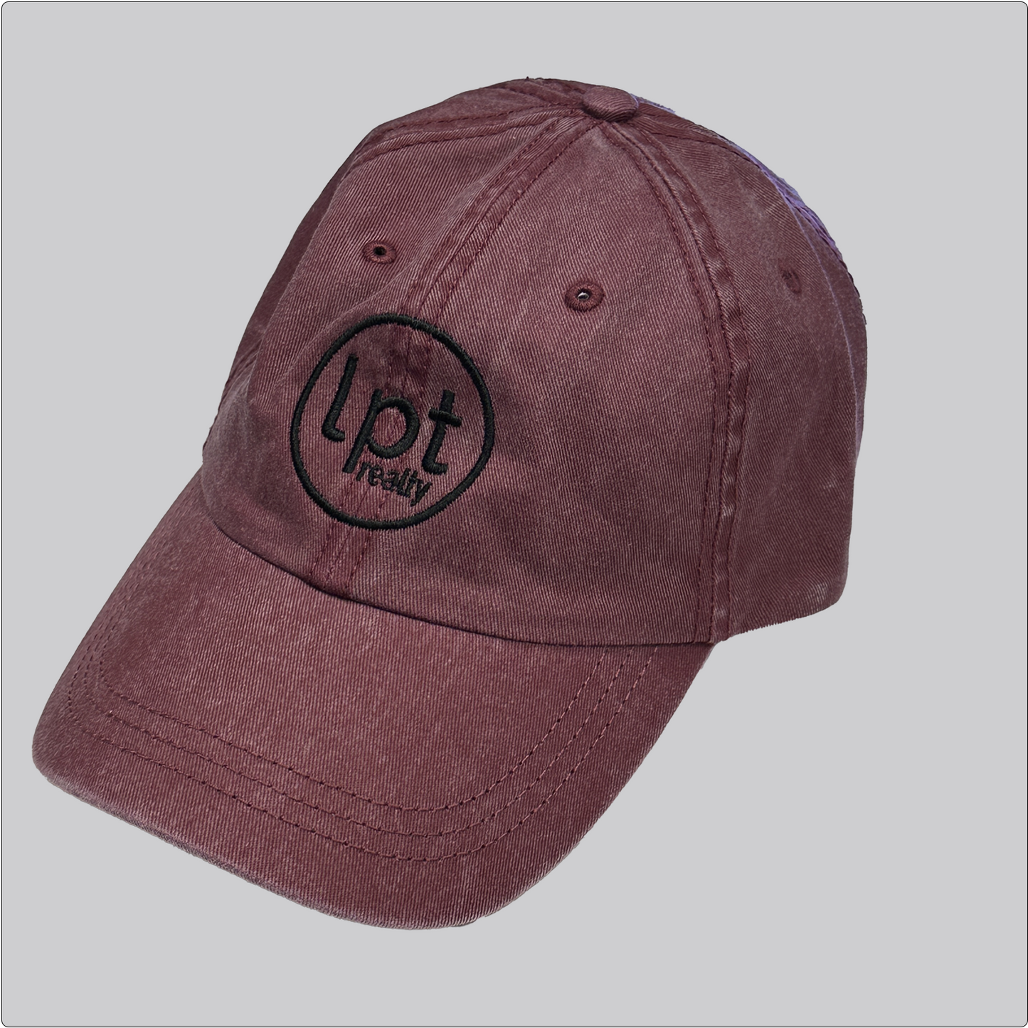 A casual, washed denim cap in a muted red with the “lpt realty” circle logo embroidered in black across the front panels. Stitching and embroidered eyelets are in matching red thread. 