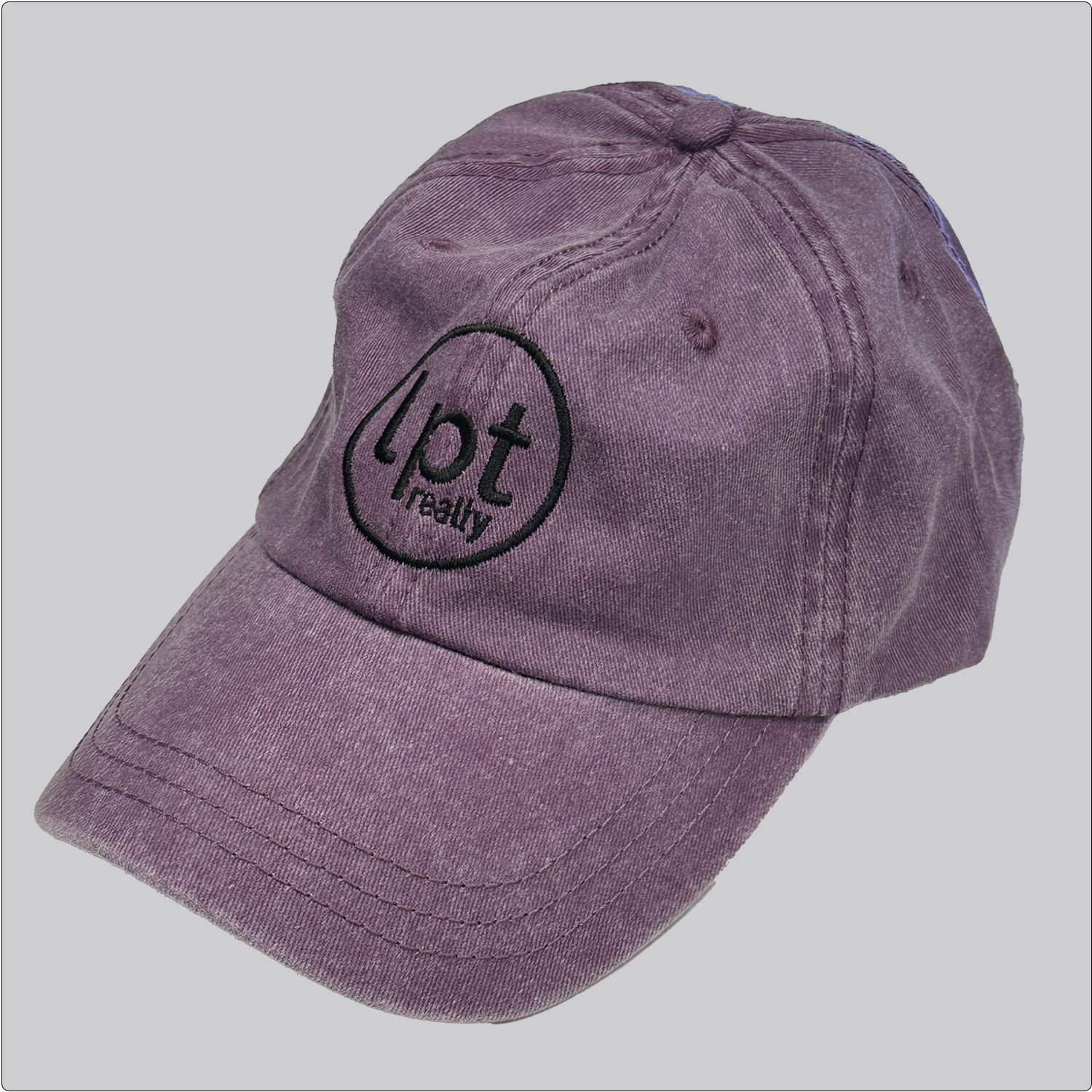 A casual, washed denim cap in a muted purple with the “lpt realty” circle logo embroidered in black across the front panels. Stitching and embroidered eyelets are in matching purple thread. 