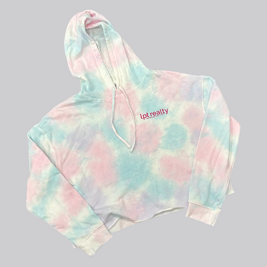 A cotton-candy blue, pink, and white tie-dyed cropped hoodie. On the top left, above the wearer’s heart, “lpt realty” is written in hot pink letters. Below that, the trademarked phrase “Brokerage For Life ™ ” appears in small, all-caps letters. The hoodie is cropped at a modest length with a raw-edge appearance on the bottom hem. On the middle-back of the hoodie, LPT’s circle logo is featured prominently in hot pink. 